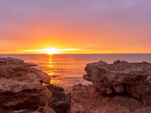 Sunset over a rocky shore with vivid orange and purple hues reflecting on the water, header image related to the blog post "A Meandering Trip Along Spain’s Unsung Orange Blossom Coast"