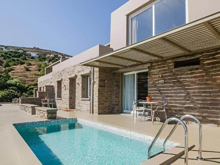 Modern house with stone walls and patio overlooking a sparkling pool. Hills in the background under a clear blue sky. Relaxed ambiance.