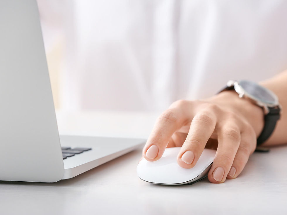 Hand using a white computer mouse next to a silver laptop on a white desk. Person wears a black watch. Bright and clean setting.