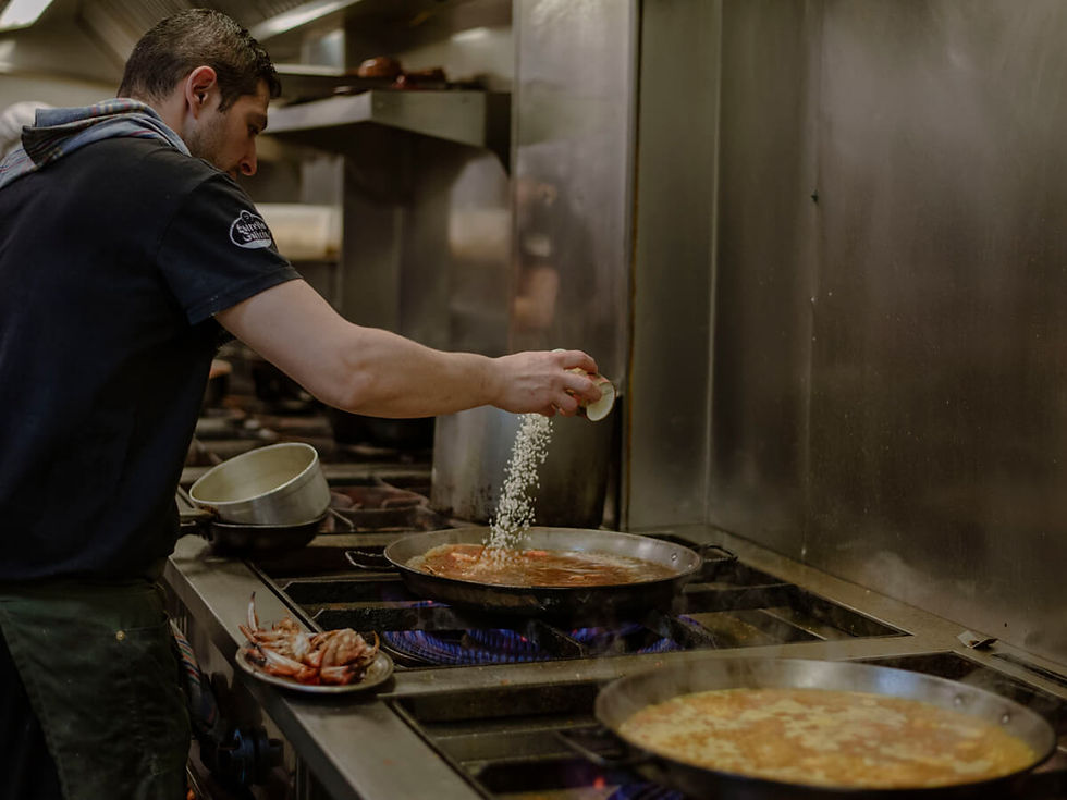 Chef adds rice to a pan in a professional kitchen. Steam rises from a stove with blue flames. A pot and crab plate are nearby.