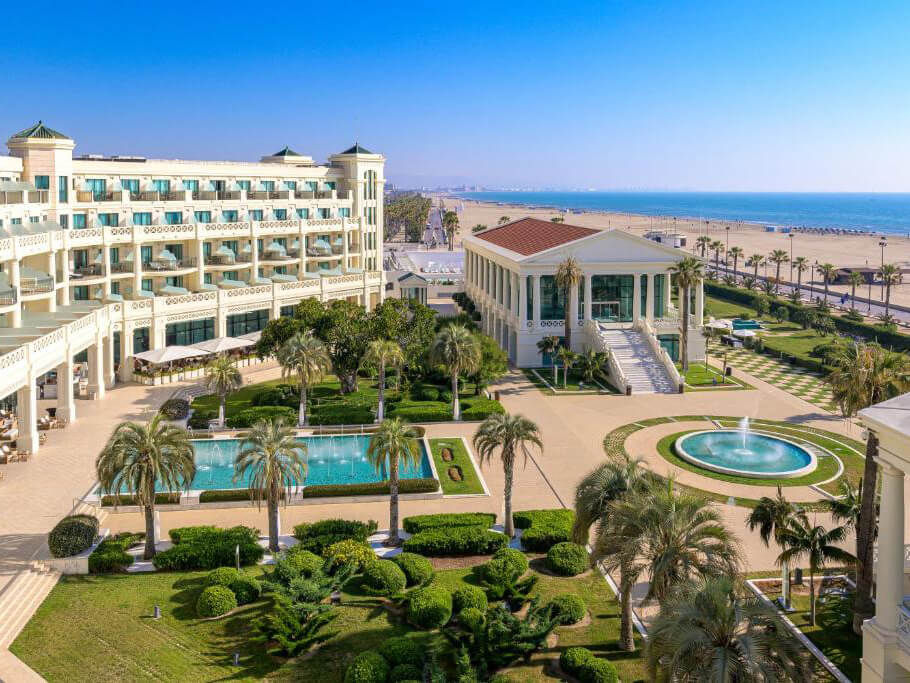 Elegant seaside hotel with lush gardens, palm trees, and fountains. Clear blue sky and beach visible, creating a serene vacation vibe.
