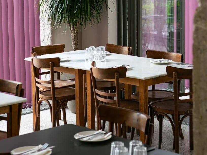 Wooden dining table with chairs, set for six. White plates and glasses on top. Pink blinds and a potted plant in the background.