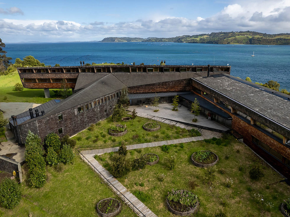 Modern building with wooden facade and garden courtyard overlooks a blue sea and green hills under a cloudy sky. Paths divide garden beds.