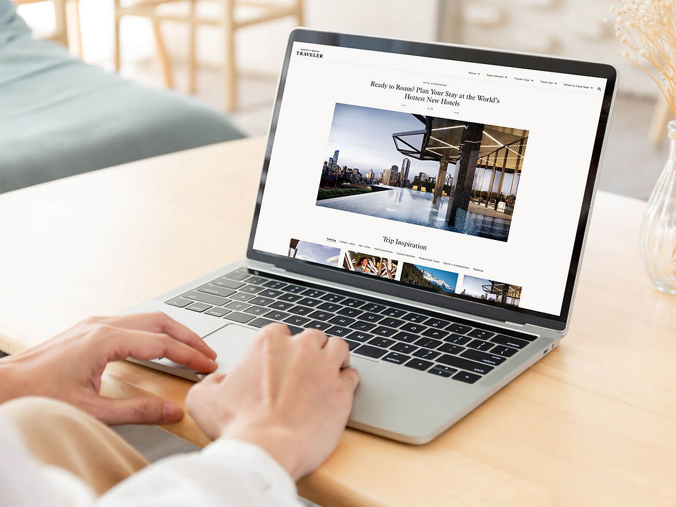 Hands typing on a laptop displaying a travel website with hotel images. The background has a wooden table and blurred furniture.