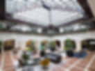 Spacious atrium with glass ceiling, modern furniture, and lush plants on rugs. Terracotta tiles and white walls create a bright, airy setting.