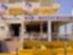 Outdoor view of "El Pescador Ermitaño" bar-restaurant with white chairs, colorful flags, and red flowers against a yellow wall. Sunny day.