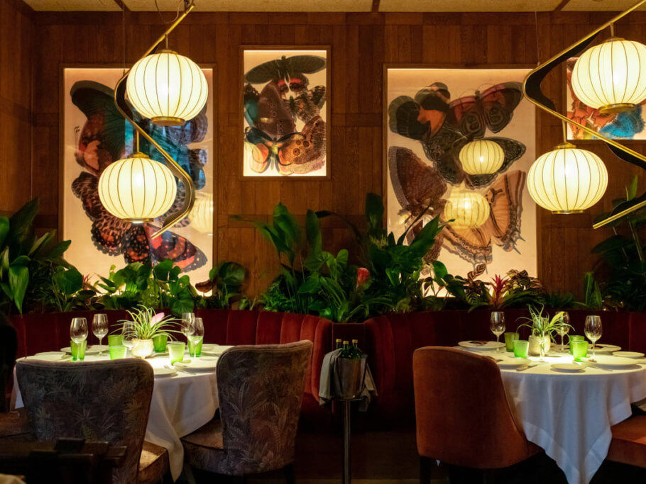 Cozy restaurant with round tables set with glasses, green plants, and butterfly art on walls. Warm lighting from hanging lamps. Relaxed mood.