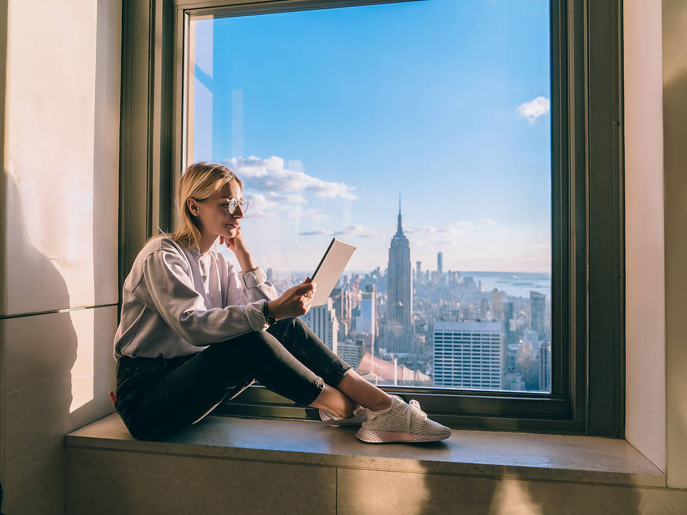 Person sitting on windowsill reading a tablet, with a sunny view of a city skyline and blue sky in the background. Calm mood.