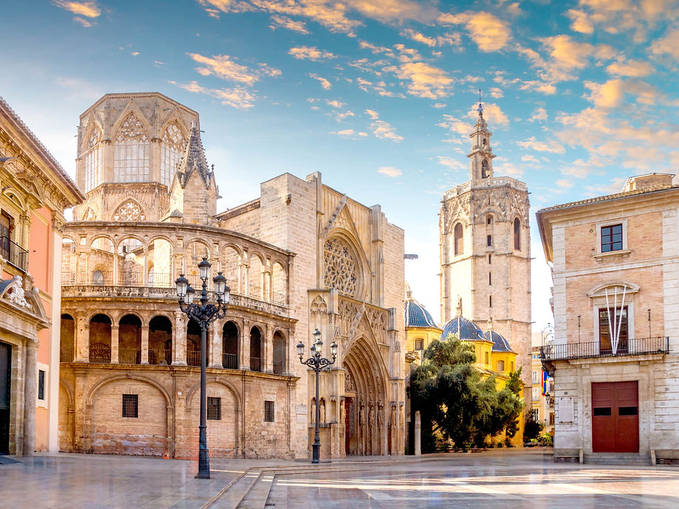 Historic cathedral and bell tower in a sunlit square, with a clear blue sky and scattered clouds. Stone buildings surround the plaza.