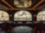 Elegant restaurant interior with a round table set with glasses and cutlery. Large arched windows reveal a sunny marina view. Warm lighting.