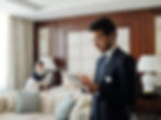 A man in a suit uses a tablet in a hotel room. A housekeeper makes the bed in the background. Neutral tones and soft lighting.