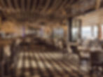 Chic cafe interior with checkered floor, rope ceiling, and tables set for dining. Large windows overlook a beach, sunny ambiance.
