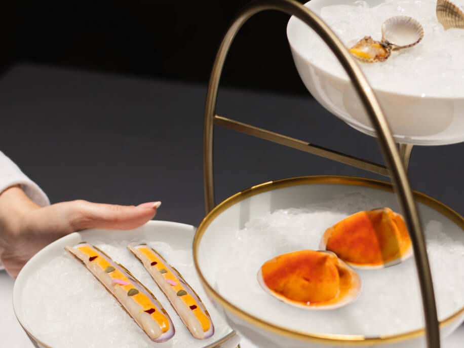 Hand reaching for seafood on a tiered platter of ice; includes orange clams and long shells with sauce, set against a dark background.