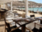 Beachside restaurant with set tables and wooden chairs under a white pergola. Sun loungers and umbrellas line the sandy shore by turquoise water.