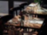 Dimly lit restaurant table set with wine glasses, menus, and flowers in a vase. Wooden chairs and tables create an elegant, cozy ambiance.