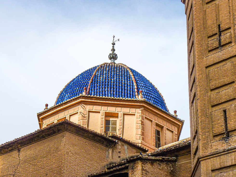 A blue-tiled dome with beige structure under a clear sky, surrounded by brown brick buildings. The scene is calm and architectural.