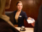 Smiling hotel receptionist in a navy blazer hands a guest a document at a wooden desk with a bell, in a warmly lit lobby.