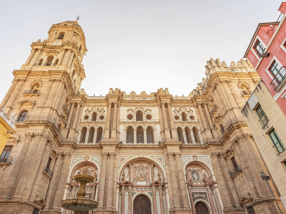 Ornate cathedral facade with tall arches and columns, set against a pale sky. A fountain is in the foreground, surrounded by colorful buildings.