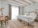 Bright bedroom with a neatly made bed, patterned pillows, wooden desk, chair, and a vase with dried plants. Soft light through curtains.