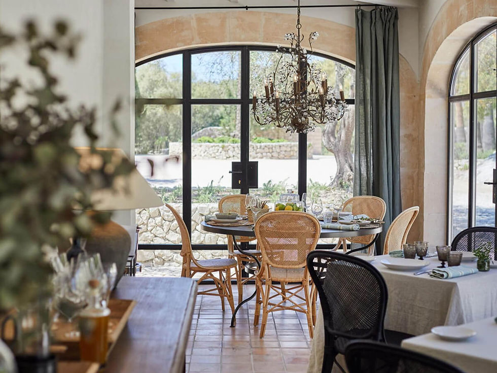 Elegant dining room with a round table set for a meal, featuring wicker chairs and a chandelier. Large windows overlook a sunny garden.