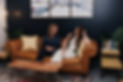 Two women laughing on a leather couch against a navy wall background.