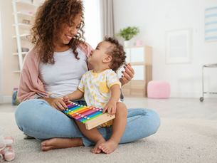 A parent sits on the floor holding a young child on their lap as they play a colourful xylophone together