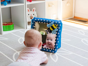 A baby lies on their tummy on a play mat, smiling at their reflection in a soft baby mirror with a small bee toy attached. Toy shelves can be seen in the background.