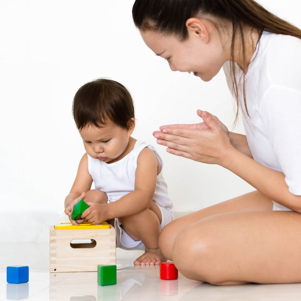 Mother claps as toddler in white onesie fits a green block into a wooden shape sorter, with red and blue blocks on a white floor.