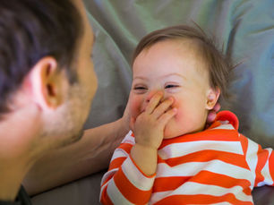 Image description: A parent smiles at a laughing young child lying on a bed. The child covers their mouth while making eye contact with the parent, sharing a playful moment together.