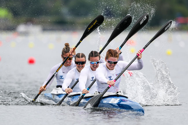 Four women in a K4 paddle in sync during a race. Emma Russell sits in seat 2, wearing white GBR kit and sunglasses.