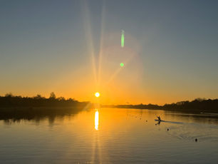 Emma Russell paddling her kayak on the right side of a calm lake at sunset, with the golden sun low on the horizon and its reflection shimmering on the water. Trees form dark silhouettes along the banks of the lake.