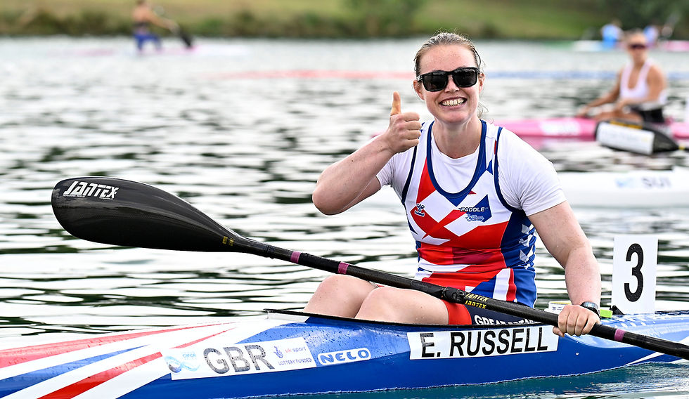 Emma Russell sits in her kayak, wearing white GBR kit with red and blue accents and sunglasses. She gives a thumbs-up and smiles, celebrating a strong race.. Other competitors are visible in the background.