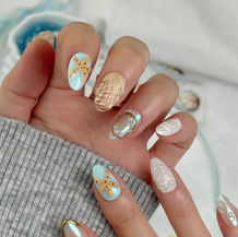 A set of medium-length almond-shaped press-on nails painted with an ocean-themed design: soft aqua and sea-blue base with glittering golden outlines. The nails feature decorative motifs including seashells, starfish, and seahorses, with shimmering gold accents that evoke a “gilded-seashell under sea” fantasy aesthetic. The nails are laid out in a fan-like arrangement against a plain background, showing their shape and detailed artwork.