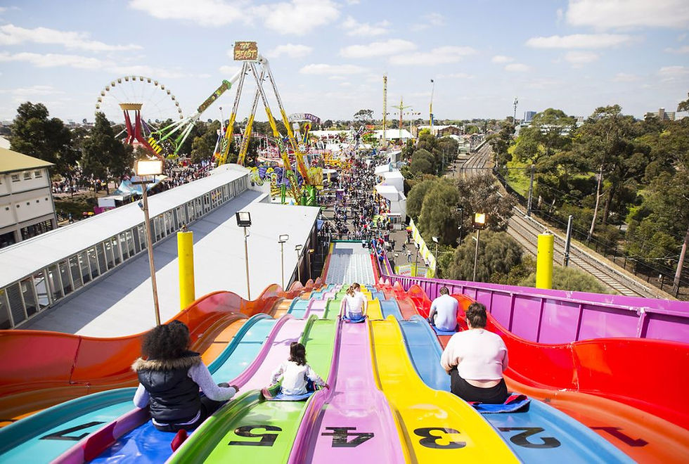 Bright carnival at sunset with colorful lights, Ferris wheel, bustling crowd, and silhouetted riders on a ride against city buildings.