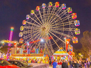 Colorful Ferris wheel lights up a busy nighttime carnival with crowds, booths, and parked cars under a dark sky.