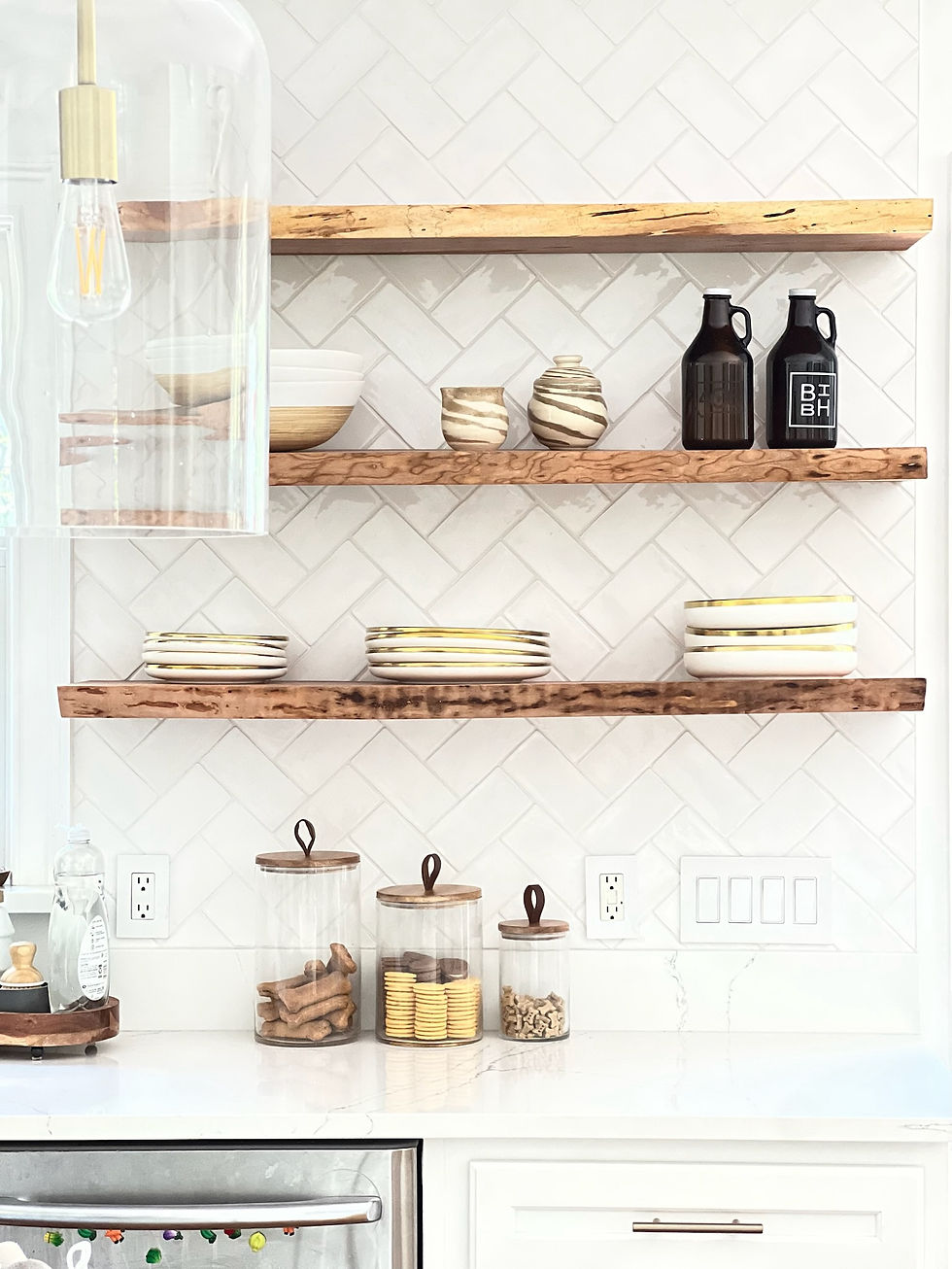 Live edge cherry floating shelves with white herringbone tile and gold-rimmed dishes in a Hamptons kitchen.