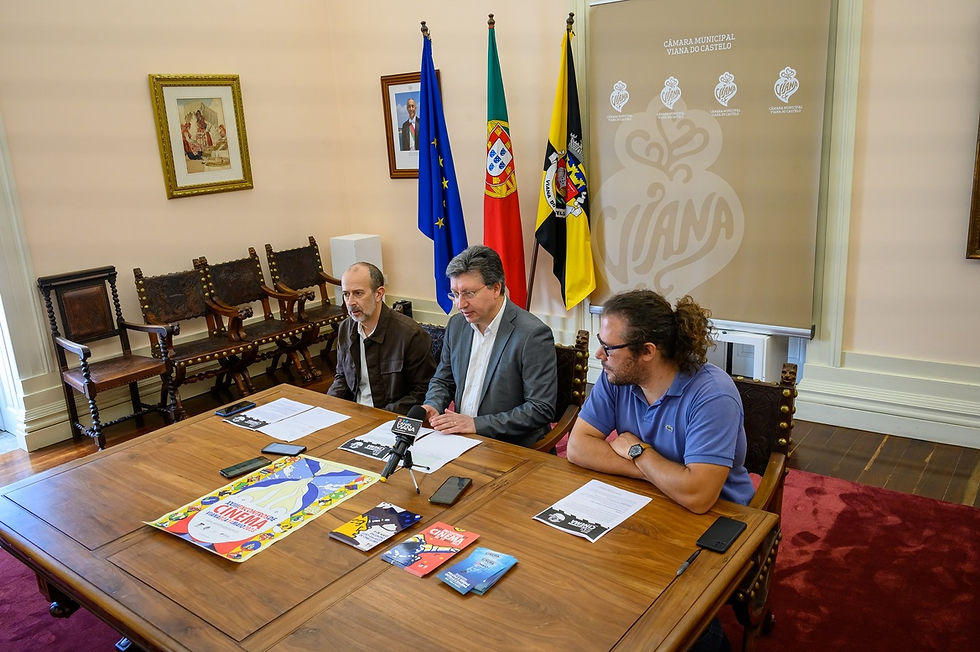 3 homens sentados a uma mesa com bandeiras atrás e microfones e telemóveis sobre a mesa