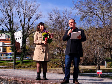 mulher de gabardine bege segurando um ramo de flores e homem a falar ao microfone