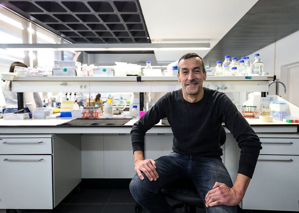 homem vestido de camisola preta e jeans, sentado, sorridente, olhando em frente. Laboratório com produtos químicos em pano de fundo.
