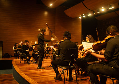 Foto lateral do maestro Andre Bachur em primeiro plano, centralizado, com a orquestra durante concerto, com mais tempo de exposição e sensação de movimento nas mãos.