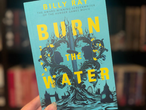 A hand holds a blue book titled "Burn the Water" by Billy Ray. The cover features ornate black designs. Blurred bookshelf in the background.