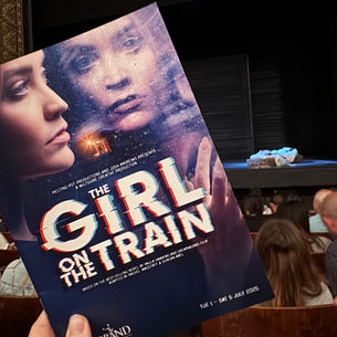 Hand holding "The Girl on the Train" program in a theater, with stage set in background. Moody cover art visible, audience seated.