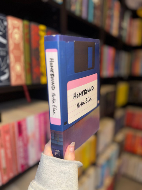 A hand holding a book whose cover is designed to look like a vintage blue floppy disk. The title Homebound and the author Portia Elan are written on a label in the centre. Shelves filled with colourful books are blurred in the background.