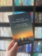 Hand holds a book with a cover showing three silhouettes against a sunset over the ocean. Text reads: "Three women. Three stages of life."