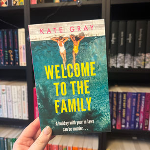 Hand holding the book "Welcome to the Family" by Kate Gray, set against colorful bookshelves. Two people in swimsuits on the cover, poolside.
