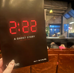 A person holds a theater program reading "2:22 A Ghost Story" in front of a stage. The setting is an ornate theater with empty seats.