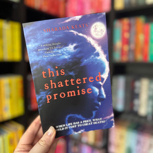 Hand holds a book titled "These Stolen Lives" by Sharada Keats. Cover shows a dramatic coastline with small red boats. Blurred bookshelf behind.