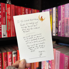 A hand holds a handwritten note in front of a colour-coordinated bookshelf with red, pink, and purple spines. The note is addressed to "The Secret Book Review" and thanks them for reading and enjoying An Old Soul. It includes a request to post the review on Amazon or Goodreads, if possible. The message is signed with a small drawing and initials.