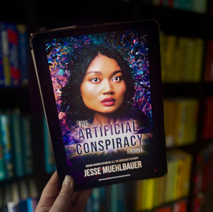 A hand holds a tablet displaying a book titled "The Artificial Conspiracy" by Jesse Muehlbauer, against a colorful, blurred bookshelf background.