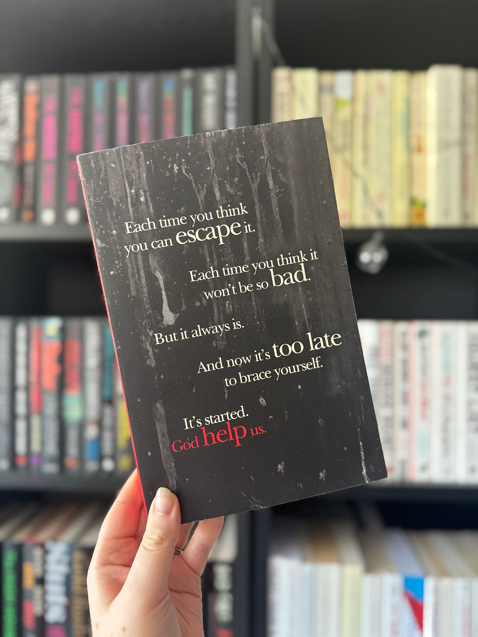 A hand holds a black book with distressed text: "Each time you think you can escape..." Bookshelves in the blurred background.